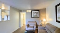 This single-floor space presents a thoughtfully arranged living area characterized by a neutral gray color palette that creates a calm and welcoming atmosphere. The living room features light wood-toned flooring that complements the soft gray walls, lending a cohesive and understated aesthetic throughout. A textured popcorn ceiling adds a layer of visual interest overhead.

The seating arrangement includes a plush upholstered armchair and a partial view of a matching sofa, both in a warm taupe tone that harmonizes with the room's color scheme. A dark coffee table anchors the seating area, while a side table with a warm-toned lamp and decorative floral accent adds a touch of softness and light to the corner of the room.

Wall décor includes a framed landscape print in a dark ornate frame, positioned prominently on the main wall. A small window near the top of the wall allows for a modest amount of natural light. An open doorway leads to an adjacent hallway or room, suggesting a connected and functional floor plan. The overall ambiance is modest, tidy, and comfortable, suited for everyday living.