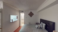 This single-floor unit offers a practical and efficiently arranged layout. The main living area combines sleeping and lounge space in an open-concept design, featuring rich hardwood flooring, crown molding, and a textured ceiling. A window dressed with draped curtains allows natural light to filter into the space, while a decorative wall piece adds a touch of character. A built-in shelving unit provides convenient storage along one wall.

The kitchen is compact yet functional, outfitted with white cabinetry offering ample storage, granite-style laminate countertops, a double-basin stainless steel sink, and a full-size refrigerator. Light tile flooring runs throughout the kitchen area, creating a clean and cohesive look.

The bathroom is well-appointed, featuring a lengthy vanity with a granite-style countertop, an undermount sink, and a wide mirror. A separate water closet provides added privacy. The tub and shower combination is finished with classic white tile and includes a curtain rod with a decorative shower curtain. Throughout the unit, a neutral white color palette creates a clean, consistent aesthetic.