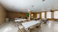 This single-floor community or event hall is designed to accommodate gatherings and group functions with practical efficiency. The main area is generously proportioned, offering ample room for multiple rectangular folding tables and chairs arranged to seat a considerable number of guests. Warm tan-toned walls give the space a neutral, welcoming character, while textured ceilings and ceiling fans distributed throughout promote comfortable air circulation. Light-toned vinyl tile flooring runs throughout, contributing to easy maintenance and a clean, open feel. Arched windows along the perimeter allow natural light to filter in, brightening the interior and creating an airy atmosphere. A decorative structural column near the center of the main room adds a subtle architectural element without interrupting the open floor plan. Additional folding chairs stored along the walls indicate the space's adaptability for varying event sizes. A support kitchen is accessible from the main hall, equipped with white cabinetry, a double sink, a full-size refrigerator, a countertop microwave, and a stovetop range — providing convenient food preparation and serving capabilities. A fire extinguisher is visibly mounted for safety compliance. Overall, this straightforward and functional space is well-suited for community events, meetings, receptions, or similar gatherings.