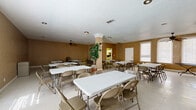 This single-floor community or event hall is designed to accommodate gatherings and group functions with practical efficiency. The main area is generously proportioned, offering ample room for multiple rectangular folding tables and chairs arranged to seat a considerable number of guests. Warm tan-toned walls give the space a neutral, welcoming character, while textured ceilings and ceiling fans distributed throughout promote comfortable air circulation. Light-toned vinyl tile flooring runs throughout, contributing to easy maintenance and a clean, open feel. Arched windows along the perimeter allow natural light to filter in, brightening the interior and creating an airy atmosphere. A decorative structural column near the center of the main room adds a subtle architectural element without interrupting the open floor plan. Additional folding chairs stored along the walls indicate the space's adaptability for varying event sizes. A support kitchen is accessible from the main hall, equipped with white cabinetry, a double sink, a full-size refrigerator, a countertop microwave, and a stovetop range — providing convenient food preparation and serving capabilities. A fire extinguisher is visibly mounted for safety compliance. Overall, this straightforward and functional space is well-suited for community events, meetings, receptions, or similar gatherings.
