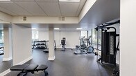 Located at 301 Green St in Portsmouth, Virginia, this fitness center offers a well-equipped exercise space spread across a single floor. The layout is open and functional, organized into two connected areas that together provide ample room for a comprehensive workout experience.

The space features a drop-panel ceiling with recessed fluorescent lighting that ensures bright, even illumination throughout. Large windows along the exterior walls invite generous natural light and offer views of the surrounding greenery and parking area, creating an airy and inviting atmosphere. White-painted structural columns punctuate the floor plan, subtly defining different workout zones without obstructing movement.

The durable rubber-speckled flooring covers the entire space, providing cushioning and traction ideal for physical activity. Equipment includes multiple cardio machines — treadmills, ellipticals, and a stationary bike — as well as a multi-station cable machine for strength training. A tiered dumbbell rack stocked with a range of free weights is positioned near the windowed wall, accompanied by an adjustable weight bench. Full-length mirrors on one wall enhance the sense of space and allow users to monitor their form. The facility also features clear exit signage and is accessible via a standard entry door, ensuring safety and convenience for all users.