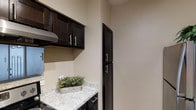 The kitchen in this single-floor space is thoughtfully designed with a cohesive aesthetic centered around dark espresso-toned cabinetry. Upper wall cabinets with sleek bar-pull hardware provide generous overhead storage, while a built-in microwave is seamlessly integrated above the range area, complemented by a ventilation hood below. A tall, narrow pantry-style cabinet tucked against the adjacent wall offers additional vertical storage, making efficient use of the available space.

The countertops feature a light granite-style surface with subtle grey and white veining, offering a striking contrast against the dark cabinetry below. The lower cabinets mirror the same hardware and finish as the uppers, creating a unified, polished look throughout. The walls are finished in a warm neutral tone, enhancing the overall brightness of the space. The layout is compact yet functional, with each element positioned to maximize both storage and workflow efficiency. The clean lines and contemporary hardware choices give the kitchen a modern, well-appointed feel suitable for everyday use.