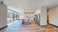 This contemporary single-floor residence showcases a clean, modern design with light wood-style flooring, crisp white walls, and teal-accented baseboards that carry consistently throughout the space. The open-concept kitchen is a standout feature, equipped with sleek white cabinetry, a subway tile backsplash, stainless steel appliances including a French door refrigerator and range, and a generous central island topped with a white quartz surface. Three stylish pendant lights hang above the island, adding warmth to the bright, airy kitchen. Floor-to-ceiling windows along one wall flood the living and kitchen areas with generous natural light and offer views of the surrounding landscape. The bedrooms are well-proportioned, each benefiting from large windows fitted with roller shades, light wood flooring, and ample closet space, including a walk-in closet outfitted with multi-tier wire shelving. The primary bathroom is finished to a high standard, featuring a dual-sink vanity with white cabinetry, stone countertops, framed mirrors, and a glass-enclosed walk-in shower with tile surround. A dedicated in-unit laundry closet with side-by-side washer and dryer adds everyday convenience. The overall ambiance is bright, clean, and thoughtfully designed for comfortable modern living.