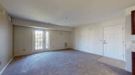 This single-floor unit offers a practical and functional layout designed to maximize livable space. The open living area is generously sized, finished with plush carpeting and neutral-toned walls, and features a set of glass-paned French doors that invite natural light and provide outdoor access. A wall-mounted heating unit adds a utilitarian touch to the space.

The kitchen is well-appointed with rich, dark wood cabinetry offering ample upper and lower storage. Stainless steel appliances include a range, microwave, dishwasher, and refrigerator, all set against light-colored countertops and durable tile flooring. The layout flows efficiently between cooking and prep zones.

The bedrooms are carpeted and bright, benefiting from multiple windows fitted with blinds. One room includes an electrical panel integrated into a structural partition wall. A walk-in closet fitted with wire shelving and hanging rods provides substantial storage capacity.

The bathroom is clean and straightforward, featuring a walk-in shower stall, a pedestal sink, tile flooring, and chrome fixtures. Overall, the unit presents a consistent, neutral aesthetic with functional finishes throughout.