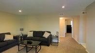This single-floor residential space offers a clean, functional layout designed for comfortable everyday living. The living room serves as the central gathering area, featuring wall-to-wall carpeting in a warm neutral tone that adds a cozy feel underfoot. The room is furnished with a dark upholstered sofa accented by decorative throw pillows, paired with a sleek glass-top coffee table and a matching side table, creating a cohesive and modern aesthetic. Recessed lighting in the ceiling provides even, ambient illumination throughout the space, complemented by a table lamp that adds a softer, warm glow. The walls are painted in a light, neutral tone, contributing to a bright and open atmosphere. A ceiling vent indicates a central climate control system for year-round comfort. A hallway visible from the living area leads to additional rooms, including what appears to be a kitchen with dark appliances, hinting at a well-connected and practical floor plan. The overall ambiance is understated and welcoming, with a straightforward design that emphasizes livability and ease of use throughout the space.