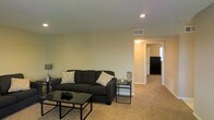 This single-floor residential space offers a clean, functional layout designed for comfortable everyday living. The living room serves as the central gathering area, featuring wall-to-wall carpeting in a warm neutral tone that adds a cozy feel underfoot. The room is furnished with a dark upholstered sofa accented by decorative throw pillows, paired with a sleek glass-top coffee table and a matching side table, creating a cohesive and modern aesthetic. Recessed lighting in the ceiling provides even, ambient illumination throughout the space, complemented by a table lamp that adds a softer, warm glow. The walls are painted in a light, neutral tone, contributing to a bright and open atmosphere. A ceiling vent indicates a central climate control system for year-round comfort. A hallway visible from the living area leads to additional rooms, including what appears to be a kitchen with dark appliances, hinting at a well-connected and practical floor plan. The overall ambiance is understated and welcoming, with a straightforward design that emphasizes livability and ease of use throughout the space.