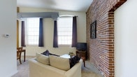 This single-floor space blends historic character with modern comfort in a compelling way. The living area serves as the centerpiece, where a full-height exposed brick wall commands attention, lending warmth and a distinct urban industrial aesthetic to the interior. The textured red brick provides a striking contrast against the smooth, neutral-toned walls and light ceiling, creating a visually dynamic environment.

Natural light pours in through large multi-pane windows dressed with dark curtain panels, illuminating the carpeted floor and brightening the overall ambiance. A wall-mounted split air conditioning unit ensures year-round climate comfort while maintaining a clean, unobtrusive look overhead.

The living area is thoughtfully arranged with a plush sofa, a central coffee table with decorative flowers, and a dedicated media console holding a flat-screen television — providing both a relaxing and functional space. Framed artwork on the brick wall adds a curated, personal touch to the room.

Overall, the space strikes a balance between rustic charm and contemporary living, with the exposed masonry as its defining architectural feature. The cozy scale and cohesive design make it an inviting and character-rich environment suited for comfortable day-to-day living.