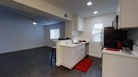 This single-story space showcases a thoughtfully designed open-concept layout that seamlessly connects the kitchen and living area. The kitchen is a standout feature, equipped with crisp white cabinetry that extends to the upper walls, providing generous storage. A central peninsula with a built-in sink and dishwasher serves as both a functional workspace and a natural divider between the kitchen and living area, with bar stool seating along one side offering a casual dining option. Stainless steel appliances, including a refrigerator and range, complement the clean aesthetic. Recessed lighting throughout the kitchen ensures bright, even illumination, while white countertops enhance the modern, airy feel. Dark hardwood flooring runs consistently across both the kitchen and living areas, creating a cohesive visual flow and striking contrast against the white walls. The living area benefits from a ceiling fan and multiple windows fitted with blinds, allowing for controlled natural light. The overall palette of white walls, white cabinetry, and dark flooring gives the space a contemporary and clean ambiance.