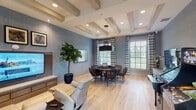 This single-floor entertainment and lounge space is thoughtfully designed to serve as a multifunctional gathering area. The room is unified by a distinctive blue textured wallcovering that spans the walls, complemented by light hardwood flooring and a coffered ceiling with exposed beams that add architectural depth and visual interest overhead. Recessed lighting throughout ensures the space is well-lit, while multiple large windows dressed with striped curtains and horizontal blinds allow generous natural light to filter in, offering views of the surrounding outdoor area.

The space is divided into functional zones. A dedicated lounge area features plush cream-colored recliner chairs arranged around a wall-mounted flat-screen television set atop a warm wood media console, flanked by framed artwork. A lush fiddle-leaf fig tree in a cobalt blue planter adds a vibrant natural element to this seating zone.

Adjacent to the lounge is a game area centered around a round poker table with built-in cup holders, surrounded by wicker-style chairs and illuminated by a striking oversized drum pendant light with a gold interior. A vintage-style arcade cabinet further enhances the entertainment offerings. Double glass doors provide direct access to the outdoors, making this a well-connected and versatile communal space.