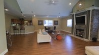 This single-floor home is designed around an expansive open-concept layout that seamlessly integrates the living, dining, and kitchen areas into one cohesive space. The living room is anchored by rich hardwood flooring that extends throughout, while soaring vaulted ceilings amplify the sense of openness and airiness. A ceiling fan with built-in lighting is centrally positioned, complementing the recessed lighting that illuminates the space evenly. Decorative wall art adds character to the neutral-toned walls, which are finished with crisp crown molding for a polished, refined look. A striking stone fireplace with a classic white mantel serves as a prominent focal point, adding warmth and texture to the room. The dining area is situated centrally within the open floor plan, offering a natural transition between the living and kitchen spaces. The kitchen features a raised breakfast bar that separates it from the main living area while maintaining visual connectivity. Multiple windows allow natural light to pour in, enhancing the bright and welcoming ambiance throughout. The thoughtful layout and combination of traditional and contemporary design elements make this a comfortable and functional living environment.