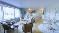 This single-floor space offers an open-concept layout that connects the living area to the kitchen, creating a cohesive and functional flow. The living room is generously proportioned, featuring warm parquet flooring underfoot and soft blue-grey walls that lend a calm, airy atmosphere throughout. Large windows along one wall invite abundant natural light, enhancing the sense of openness. The seating area is centered around a light-toned sofa complemented by accent chairs and layered area rugs, offering a comfortable and well-appointed gathering space.

The kitchen is positioned at the rear of the open-plan area, featuring a raised breakfast bar that serves as a natural divider between the living and cooking spaces. It is outfitted with contemporary cabinetry in a muted tone, stainless steel appliances including a built-in microwave, and dark stone countertops that add contrast to the overall palette. A hallway visible from the main space leads to additional rooms, with a wall-mounted light fixture providing a welcoming transition. The overall ambiance is bright, cohesive, and thoughtfully arranged.