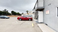This surface parking lot is situated directly alongside a multi-story building and offers a generous open-air parking area with clearly marked stalls and yellow wheel stops throughout. The paved lot accommodates multiple vehicles with ease, providing straightforward access for both residents and visitors. A metal exterior staircase connects the ground level to the upper floor of the adjacent building, positioned near a ground-level entry door for convenient access. The surrounding environment features a mix of low-rise commercial and residential structures, mature trees, and a palm tree visible in the background, lending a California character to the setting. The lot itself is fully exposed to the elements, with no overhead covering, allowing for an open and unobstructed environment. The building adjacent to the lot features a second-floor balcony with metal railings, visible from the parking area. The overall setting is functional and practical, designed to prioritize vehicle access and ease of movement, with the surrounding streetscape and greenery providing a modest sense of context and place.