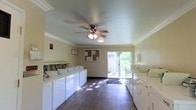 This shared laundry facility is a spacious, single-floor room designed for practical, everyday use. Rows of white top-load washers and dryers line both sides of the room, providing ample capacity for multiple users simultaneously. The flooring consists of rich, dark hardwood-style planks that contrast cleanly with the light-colored walls, lending the space a tidy and well-maintained appearance.

Crown molding runs along the perimeter of the ceiling, adding a subtle architectural detail that elevates the room's overall finish. A ceiling fan with an integrated light fixture is centrally mounted, promoting air circulation and ensuring the space remains comfortable during use.

Natural light is a notable feature of this room, streaming in through a large sliding glass door at the far end that opens to a lush, green outdoor area. A window above the door further enhances the brightness of the interior. A cork bulletin board is mounted on the wall near the entrance, serving as a practical information station for notices and announcements.

The room also includes a wall-mounted utility shelf area above the washers, offering convenient space for laundry supplies. Overall, the facility is clean, functional, and thoughtfully appointed, making it a practical and comfortable space for shared laundry needs.
