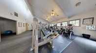 Located at 2931 Cottage Pl in Greensboro, NC, this fitness center offers a thoughtfully designed workout facility across a single floor. The space is defined by its soaring vaulted ceilings with exposed beam detailing, which lend an open, airy atmosphere throughout. White walls and multiple windows along the perimeter allow abundant natural light to fill the room, creating an inviting and energizing environment.

The main workout area is outfitted with a robust selection of professional-grade equipment. Strength training is well-represented with multiple Hoist selectorized cable and resistance machines, a power rack with weight plates and battle ropes, a dumbbell rack, and dedicated benches. For cardiovascular training, the facility includes treadmills, elliptical trainers, and a rowing machine. A wall-mounted flat-screen television adds to the overall comfort of the space.

Rubber flooring covers the primary equipment zones, providing safety and durability, while carpet covers additional floor areas. Framed informational posters are displayed on the walls, and elegant chandelier-style ceiling fixtures add a refined touch. A separate adjoining room, visible through an interior doorway, suggests additional functional space. The facility also features a clearly marked emergency exit and proper safety signage, reflecting a well-maintained and professional environment.