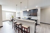 Located at 1325 Sunset Lake Dr in Shiloh, IL, this single-floor home offers a thoughtfully designed layout across a generous footprint. The open-concept living area seamlessly connects the kitchen, dining room, and living room, creating an inviting flow throughout. The kitchen is a standout, showcasing dark espresso cabinetry, granite countertops, a classic subway tile backsplash, stainless steel appliances, and a large island with bar seating illuminated by stylish pendant lighting. The adjacent dining room features a chandelier and natural light from surrounding windows. The living room offers a cozy fireplace and light wood-style flooring that extends throughout the main areas. A wide hallway connects the private quarters, where the spacious primary bedroom features vaulted ceilings, a ceiling fan, and ample natural light. The primary bath includes a walk-in tile shower with a handheld fixture and grab bar. Two well-sized walk-in closets provide generous storage. Three bathrooms in total serve the home. A dedicated laundry room houses a full-size washer and dryer with overhead shelving. The home's neutral palette and modern finishes create a clean, comfortable, and functional living environment throughout.