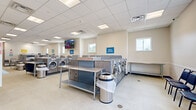 This commercial laundromat is a clean, efficiently designed single-floor facility built to accommodate a high volume of customers. The open floor plan is organized to maximize machine accessibility, featuring rows of front-loading washers in multiple load capacities — including 20 lb, 40 lb, and 90 lb machines — clearly labeled with prominent signage to help customers quickly find the right equipment for their needs. A bank of stacked 30 lb dryers lines one wall, offering efficient drying options alongside the washers. Stainless steel folding tables are centrally positioned throughout the space, providing ample workspace for sorting and folding laundry. The facility includes a dedicated soap center stocked with soap, bleach, softeners, and laundry bags, as well as a bill and coin changer for added convenience. Wall-mounted flat-screen televisions are positioned throughout the facility, allowing customers to stay entertained during their visit. Seating is available for those waiting. The interior features a drop-panel ceiling with recessed lighting, light-toned flooring, and windows that bring in natural light. The entrance area is well-marked with posted guidelines and an access-controlled door, contributing to an organized and secure environment overall.