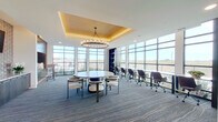 This single-floor workspace delivers a polished, professional environment designed to accommodate both collaborative gatherings and focused individual work. The room's most striking architectural feature is a recessed tray ceiling finished in warm gold tones, from which a large circular chandelier with exposed Edison-style bulbs is suspended, creating an elegant focal point above the central meeting area.

Floor-to-ceiling windows wrap along multiple walls, flooding the interior with natural light and offering unobstructed views of the surrounding landscape. The patterned carpet in cool blue-grey tones with subtle wave detailing anchors the space and adds visual texture underfoot.

The layout is divided into two functional zones. The primary area centers on a large round white table surrounded by a mix of upholstered lounge chairs, ideal for collaborative discussions. Adjacent to this, a long workstation with ergonomic rolling chairs lines the windowed wall, providing a dedicated workspace for individual tasks.

A full-height dark-toned built-in shelving unit displays decorative objects and adds depth along one wall. A separate lounge nook features a cushioned banquette, high-top table, and bar-height seating set against a rich wood-paneled accent wall, offering a more relaxed setting within the same open-plan environment.