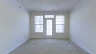 This single-floor space offers a clean, understated aesthetic defined by its light-filled interior and neutral color palette. The main living area is finished with plush, light beige wall-to-wall carpeting that adds warmth underfoot, while the crisp white walls and crown molding provide a polished, classic feel throughout.

Natural light is a standout feature of this space. A central glass-panel exterior door is flanked by two large double-hung windows, each fitted with horizontal blinds for adjustable light control. Together, these openings flood the interior with bright, even daylight and offer views of the surrounding greenery outside. The transom window above the door further enhances the sense of openness and allows additional light to filter in.

The ceiling is smooth and finished in white, contributing to the room's airy atmosphere. Recessed ceiling vents indicate a central HVAC system for year-round climate comfort. The overall layout is open and versatile, providing a blank canvas well-suited for a variety of furniture arrangements and functional uses. The space presents a move-in-ready environment with a straightforward, practical design.