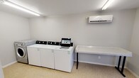 Located at 1025 W Wheatland Rd in Dallas, Texas, this single-floor commercial laundry facility is designed to provide a functional and efficient laundry experience. The space is divided into two connected areas: a main laundry room and an adjacent waiting area.

The main laundry room is equipped with multiple top-load washing machines, including a digital payment-enabled unit, as well as stacked front-load dryer units, offering ample capacity for high-volume use. A stainless steel front-load washer and dryer set rounds out the appliance offerings. A wall-mounted mini-split air conditioning unit ensures climate comfort throughout the room. A sturdy folding table with a speckled countertop surface provides a dedicated space for sorting and folding laundry. The room is lit by fluorescent ceiling fixtures and features neutral gray walls, textured ceilings, and durable speckled flooring throughout.

The adjoining waiting area offers seating for guests, with a row of molded plastic chairs in alternating black and white, connected by a shared armrest frame. This area is similarly well-lit and monitored by a security camera. A direct exterior exit door provides convenient access to the outdoor surroundings, including a covered walkway and parking area. The overall design is clean, utilitarian, and well-maintained, making this facility a practical resource for its users.