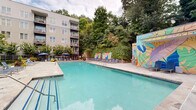 Located at 390 Stovall St SE in Atlanta, Georgia, this outdoor amenity space offers a well-appointed pool area designed for both relaxation and social gathering. The centerpiece is a generously sized swimming pool with clear turquoise water, surrounded by an expansive stone-paved deck finished in natural flagstone. Ample blue lounge chairs and umbrella-shaded seating are arranged throughout the deck, providing plenty of space for sunbathing and leisure.

A defining artistic feature of this space is a series of bold, colorful murals depicting tropical botanical motifs in vivid blues, greens, and pinks. These murals are displayed along a rustic stone retaining wall draped in lush ivy, adding a vibrant and creative backdrop to the pool environment. Mature trees border the perimeter, offering natural shade and a sense of privacy.

Adjacent to the pool, a dedicated outdoor entertaining area includes a built-in grill station with stone surround, along with dining tables and chairs set beneath a covered canopy. This space is ideal for outdoor cooking and communal dining. The multi-story residential building frames the courtyard on multiple sides, with private balconies overlooking the amenity area below. The overall ambiance is lively, colorful, and inviting, blending natural greenery with artistic design elements.