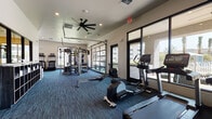 This fitness center is a thoughtfully designed, single-floor facility offering a comprehensive range of workout equipment and an energizing atmosphere. The open floor plan is covered in durable, multi-toned blue carpet tiles, and the space is illuminated by recessed lighting complemented by ceiling fans that promote airflow throughout.

The cardio zone is well-appointed with multiple Life Fitness treadmills, elliptical trainers, and a rowing machine, all positioned to take advantage of the large floor-to-ceiling windows that offer views of the outdoor amenity area. A wall-mounted flat-screen TV accompanies this section, adding an entertainment option during workouts.

A dramatic motivational mural — depicting a silhouetted figure helping another reach a summit against a vivid sunset sky — serves as a bold focal point and adds visual energy to the space. Nearby, a dedicated strength and functional training area features a Life Fitness multi-functional cable and rack system, along with a JumpSport trampoline and additional accessories to support a variety of training styles.

Built-in cubby shelving along one wall provides organized storage for towels and accessories. Large glass garage-style doors and expansive windows flood the space with natural light and create a seamless visual connection to the outdoors, enhancing the overall openness and motivation of the environment.