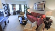 This single-floor residence centers around a thoughtfully designed living room that balances comfort and style. The space is anchored by a rich red sofa adorned with dark accent pillows, complemented by striped armchairs that add a classic, layered aesthetic. A round dark-wood coffee table serves as a focal point, offering both functionality and visual grounding to the seating arrangement. Neutral wall-to-wall carpeting runs throughout, contributing to the warm and inviting ambiance of the room. Natural light filters in through large windows dressed with elegant floor-length draping curtains in a muted green tone, enhancing the sense of openness. A tall indoor plant adds a refreshing organic element to the corner of the room. The walls are kept in a soft, neutral tone, providing a clean backdrop that allows the furnishings and artwork to stand out — notably, a framed botanical print displayed prominently above the sofa. In the background, a dedicated area with a desk and task lamp suggests a functional workspace or study nook, making the space both livable and practical. Overall, the layout feels spacious, cohesive, and move-in ready.