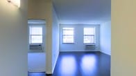 This single-floor space presents a straightforward yet functional layout characterized by clean lines and an open feel. The interior is defined by crisp white walls that enhance the sense of brightness throughout, complemented by dark hardwood floors that add contrast and a polished finish to the space. The flooring reflects natural light effectively, contributing to the overall luminosity of the interior.

Multiple double-hung windows are positioned along the exterior wall, allowing generous amounts of natural light to filter in and illuminate the space. Beneath each window, wall-mounted HVAC units provide climate control, a practical feature integrated seamlessly into the design. The windows offer views of the surrounding urban environment, reinforcing the city context of the space.

An open doorway connects adjacent areas, suggesting a multi-room configuration that allows for flexible use. The walls transition smoothly between rooms, maintaining a cohesive aesthetic throughout. The absence of ornamental detailing keeps the interior neutral and adaptable, making it well-suited for a variety of uses. Overall, the space conveys a clean, minimalist character with functional amenities in place.