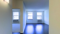 This single-floor space presents a straightforward yet functional layout characterized by clean lines and an open feel. The interior is defined by crisp white walls that enhance the sense of brightness throughout, complemented by dark hardwood floors that add contrast and a polished finish to the space. The flooring reflects natural light effectively, contributing to the overall luminosity of the interior.

Multiple double-hung windows are positioned along the exterior wall, allowing generous amounts of natural light to filter in and illuminate the space. Beneath each window, wall-mounted HVAC units provide climate control, a practical feature integrated seamlessly into the design. The windows offer views of the surrounding urban environment, reinforcing the city context of the space.

An open doorway connects adjacent areas, suggesting a multi-room configuration that allows for flexible use. The walls transition smoothly between rooms, maintaining a cohesive aesthetic throughout. The absence of ornamental detailing keeps the interior neutral and adaptable, making it well-suited for a variety of uses. Overall, the space conveys a clean, minimalist character with functional amenities in place.