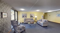 This single-floor space centers around a thoughtfully designed living area that combines comfort and coastal charm. The room is painted in a warm, soft yellow that lends an inviting and cheerful ambiance throughout. Underfoot, deep blue carpeting complements the color palette and adds a layer of coziness to the space.

Seating is generously provided through a combination of two neutral-toned sofas and a pair of boldly patterned armchairs in blue and white ikat fabric, creating a welcoming arrangement ideal for relaxed gatherings. A round glass-top coffee table with an ornate metal base serves as a central focal point, styled with decorative accents that reinforce the coastal theme.

Two framed landscape prints depicting beach and dune scenes hang on the yellow accent wall, enhancing the seaside aesthetic. A traditional floor lamp provides warm ambient lighting alongside flush-mounted ceiling fixtures. Large windows along one wall allow natural light to filter into the space.

The textured ceiling adds subtle architectural interest, while a partially visible corridor hints at additional areas beyond the main living room, suggesting a functional and well-organized layout overall.