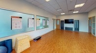 This single-floor fitness studio is designed to support a wide range of exercise activities in a clean, organized environment. The main workout area is anchored by warm hardwood flooring that extends throughout the open space, providing a durable and visually inviting surface for various training modalities. A full-length wall mirror runs along one side of the room, enhancing the sense of space while allowing users to monitor their form during exercise.

The studio features a drop ceiling with recessed lighting panels that provide consistent, even illumination throughout the room. Large interior windows along one wall display workout charts and training guides, offering easy reference for participants and instructors alike. A wall-mounted flat-screen television is positioned near one end of the room, suitable for instructional content or guided workouts.

Equipment visible in the space includes a plyo box, an aerobic step platform, a stability ball, free weights, a weight rack, and a workout bench, supporting both strength and cardio-based training. Built-in storage units and shelving along the walls help keep equipment organized. A locker area near the entrance adds practical functionality. The overall ambiance is bright, professional, and purpose-built for fitness use.