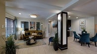 This well-appointed single-floor space showcases a thoughtfully designed open-concept layout that seamlessly connects the living area to the kitchenette. The living room is anchored by a plush olive-green sofa accented with decorative navy blue pillows, complemented by a sleek glass coffee table and a patterned area rug. Coastal-themed artwork, including a sailing scene, adorns the walls and contributes to the space's relaxed yet refined atmosphere. A striking dark-wood decorative column with integrated lighting serves as an elegant architectural divider, adding visual interest and defining the transition between zones. The kitchenette is outfitted with dark cabinetry, stainless steel appliances, and a compact, functional layout. Light-toned large-format tile flooring runs throughout, enhancing the sense of openness and reflecting the ambient lighting effectively. Recessed lighting and flush-mount ceiling fixtures provide warm, even illumination across the space. Neutral walls with subtle textured wallcovering accents create a sophisticated backdrop. A lush potted plant near the column adds a natural, organic element that softens the overall aesthetic. The space presents a cohesive blend of coastal charm and contemporary design.