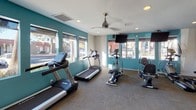 This single-floor fitness center is designed to support a comprehensive workout experience in a bright, welcoming environment. The space is divided into distinct functional zones, each tailored to a specific type of exercise. Bold teal accent walls provide a vibrant contrast to the white ceilings and neutral carpeted flooring, creating an energetic atmosphere that encourages activity.

The cardio zone is lined with large windows that flood the area with natural light and offer views of the surrounding outdoor landscape, including trees and open sky. Treadmills and an elliptical machine are positioned to take full advantage of these views. Ceiling fans overhead ensure comfortable airflow throughout workouts.

Transitioning into the strength training area, the flooring shifts to durable rubber matting, providing safety and stability. This section houses a multi-station cable machine, a power tower for bodyweight exercises, a set of dumbbells and medicine balls, and a rowing machine. A wall of mirrors expands the sense of space and allows users to monitor their form. A motivational wall decal reading "The Body Achieves What the Mind Believes" adds an inspiring touch to the atmosphere.

Recessed lighting throughout ensures the space remains well-lit at all times, complementing the abundant natural light. The overall layout is open and functional, making efficient use of the available floor space.