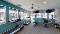 This single-floor fitness center is designed to support a comprehensive workout experience in a bright, welcoming environment. The space is divided into distinct functional zones, each tailored to a specific type of exercise. Bold teal accent walls provide a vibrant contrast to the white ceilings and neutral carpeted flooring, creating an energetic atmosphere that encourages activity.

The cardio zone is lined with large windows that flood the area with natural light and offer views of the surrounding outdoor landscape, including trees and open sky. Treadmills and an elliptical machine are positioned to take full advantage of these views. Ceiling fans overhead ensure comfortable airflow throughout workouts.

Transitioning into the strength training area, the flooring shifts to durable rubber matting, providing safety and stability. This section houses a multi-station cable machine, a power tower for bodyweight exercises, a set of dumbbells and medicine balls, and a rowing machine. A wall of mirrors expands the sense of space and allows users to monitor their form. A motivational wall decal reading "The Body Achieves What the Mind Believes" adds an inspiring touch to the atmosphere.

Recessed lighting throughout ensures the space remains well-lit at all times, complementing the abundant natural light. The overall layout is open and functional, making efficient use of the available floor space.