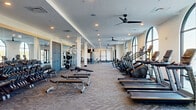 This expansive single-level fitness center is thoughtfully designed to accommodate a wide range of workout needs. The space is characterized by a series of striking arched floor-to-ceiling windows along one wall, flooding the interior with natural light and offering views of the surrounding landscape. Ceiling fans are positioned throughout, complementing the recessed lighting overhead to maintain a comfortable environment.

The main floor is divided into clearly defined workout zones. A dedicated cardio section lines the windowed wall, housing a comprehensive row of treadmills, elliptical trainers, stair climbers, stationary bikes, and recumbent cycles, many equipped with individual screens. Adjacent to this is a well-stocked strength training area featuring free weights, adjustable benches, a full dumbbell rack, and multiple cable and resistance machines. Large wall mirrors enhance the sense of space in this zone.

A separate alcove houses a power rack with barbell and weight plates set atop a protective rubber mat, ideal for heavy compound lifts. A distinct studio space, accessible through glass-paneled sliding doors and accented by a modern chandelier, provides a dedicated area for stretching, yoga, or group classes. Patterned carpet flooring ties the entire facility together with a polished, contemporary aesthetic.