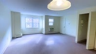 This single-floor space presents an open, versatile room finished with wall-to-wall carpeting in a neutral tone, contributing to a clean and functional aesthetic. The room is illuminated naturally by multiple windows, including a large double-pane window along one wall and a smaller window centered on an adjacent wall, allowing daylight to fill the interior and create a bright, airy atmosphere. A centrally mounted dome ceiling light fixture provides additional overhead lighting. Climate control is handled by baseboard heating units running along the base of the walls, supplemented by a wall-mounted air conditioning unit beneath one of the windows, ensuring year-round comfort. The walls are finished in a soft off-white, keeping the space feeling open and neutral. A door on the far right wall provides access to an adjoining area, suggesting additional rooms or utility spaces beyond. The overall layout is straightforward and unobstructed, offering flexibility for various uses. The space maintains a practical, no-frills character suited to functional day-to-day living or occupancy.