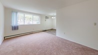 This single-floor 1-bedroom, 1-bathroom unit at Hazelwood in Inkster, Michigan presents a clean, well-organized layout designed for comfortable everyday living. The living area features soft wall-to-wall carpeting underfoot and a large picture window that fills the space with natural light and provides views of the surrounding greenery. A wall-mounted air conditioning unit and baseboard heating ensure year-round climate comfort. Adjacent to the living area, a defined dining space is accented by a classic chandelier and durable wood-look flooring that continues into the kitchen. The galley-style kitchen is efficiently arranged with rich dark wood cabinetry lining both sides, white countertops, a built-in sink, and white appliances including a coil-burner range and refrigerator, providing ample storage and prep space. The bedroom is a generously sized, quiet retreat with plush carpeting, a wide window overlooking outdoor scenery, and baseboard heating. A spacious walk-in closet off the bedroom offers dual shelving on both sides, providing substantial storage capacity. The full bathroom features classic white tile surrounding a full soaking tub and shower, a pedestal-style vanity with a mirror cabinet, and mosaic tile flooring, delivering a clean and functional design throughout.