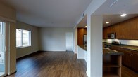 This single-floor residence offers a thoughtfully designed open-concept layout that seamlessly connects the living and kitchen areas. Rich, dark hardwood flooring extends throughout the main living space, adding warmth and a sense of continuity to the interior. The living area is generously proportioned, bathed in natural light from a window along the exterior wall, and features smooth white walls and ceilings that contribute to a clean, airy atmosphere. Subtle architectural details, including a structural column and defined half-wall partitions, elegantly delineate the living space from the kitchen without sacrificing the open feel. The kitchen is well-appointed, showcasing warm medium-toned wood cabinetry that rises toward the ceiling, providing ample storage. Stainless steel appliances, including a refrigerator and range, complement the granite countertops, lending a modern and functional character to the culinary space. Recessed lighting in the kitchen ensures the area is well-illuminated. A hallway visible from the main living area leads to additional rooms, suggesting a practical and well-organized floor plan. Overall, the space strikes a balance between modern finishes and warm, inviting design elements.