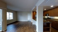 This single-floor residence offers a thoughtfully designed open-concept layout that seamlessly connects the living and kitchen areas. Rich, dark hardwood flooring extends throughout the main living space, adding warmth and a sense of continuity to the interior. The living area is generously proportioned, bathed in natural light from a window along the exterior wall, and features smooth white walls and ceilings that contribute to a clean, airy atmosphere. Subtle architectural details, including a structural column and defined half-wall partitions, elegantly delineate the living space from the kitchen without sacrificing the open feel. The kitchen is well-appointed, showcasing warm medium-toned wood cabinetry that rises toward the ceiling, providing ample storage. Stainless steel appliances, including a refrigerator and range, complement the granite countertops, lending a modern and functional character to the culinary space. Recessed lighting in the kitchen ensures the area is well-illuminated. A hallway visible from the main living area leads to additional rooms, suggesting a practical and well-organized floor plan. Overall, the space strikes a balance between modern finishes and warm, inviting design elements.