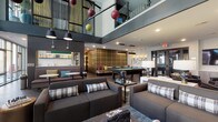 Located in the heart of Columbus, Ohio, the Nicholas Luxury Living amenity lounge at 12 W Gay St is a thoughtfully designed communal space that blends modern sophistication with vibrant, social energy. The open-concept layout soars with dramatic double-height ceilings, an open mezzanine level with glass railings overlooking the main floor, and an abundance of natural light streaming through floor-to-ceiling windows along the perimeter.

The main lounge area features plush, dark upholstered sofas accented with plaid throw pillows, arranged around low coffee tables to create inviting conversation zones. A full-size billiards table anchors the game area, set against a bold plaid-paneled accent wall and illuminated by pendant lighting. Adjacent to the game area, a wet bar with backlit shelving and counter seating provides a stylish social hub. A communal dining table surrounded by teal leather chairs offers a dedicated space for group gatherings.

A standout metallic tile feature wall provides a striking industrial backdrop for dual wall-mounted flat-screen televisions in a dedicated media lounge. A separate, more intimate screening and sitting room showcases tufted black leather armchairs, a vibrant fuchsia area rug, curated wall art, and a large-screen display — ideal for private viewings. The space is finished with warm hardwood-style flooring, designer lighting, and eclectic decor throughout.