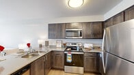 This well-appointed one-bedroom, one-bathroom unit in Spokane Valley, Washington offers a thoughtfully designed layout that maximizes comfort and functionality. The open-concept kitchen is a standout feature, outfitted with rich dark-wood cabinetry, stainless steel appliances including a refrigerator, range, microwave, and dishwasher, complemented by light countertops and easy-care hardwood-style flooring. A peninsula with seating provides a convenient dining option and enhances the kitchen's open feel. The living room is filled with natural light streaming through large windows and a glass-paneled door that opens to a private balcony, offering outdoor views and a bright, airy atmosphere. Soft carpeting throughout the living and bedroom areas adds warmth underfoot. The bedroom is a comfortable retreat, featuring a ceiling fan, a tray ceiling detail, a generous closet, and a window dressed with vertical blinds. The bathroom is efficiently designed with dark cabinetry matching the kitchen, a well-lit vanity mirror, and a convenient in-unit stacked washer and dryer tucked neatly within a closet. Neutral tones and clean finishes throughout the unit create a cohesive, modern aesthetic suited for easy living.