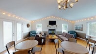 Located at 1770 Tanglewood Ct in Burlington, Kentucky, this single-floor gathering space offers a warm and versatile environment well-suited for events, meetings, or social functions. The open layout is anchored by soaring vaulted ceilings that lend an airy sense of volume to the main area, which is lined with globe string lights that trace the roofline and contribute to the space's welcoming ambiance.

The main gathering area features a central fireplace with a classic white mantel and wall-mounted television, flanked by multiple windows that allow natural light to fill the room. Warm hardwood flooring extends throughout, tying together distinct seating and dining zones. Plush sofas, armchairs, and round tables with chairs are thoughtfully arranged to accommodate both casual lounging and seated gatherings.

A functional kitchen is equipped with stainless steel appliances, including a full-size refrigerator and range, white cabinetry, and pendant lighting—ideal for event catering needs. A dedicated flex room features a large wall mirror, a folding table, and string lighting, offering additional utility space. Etched-glass French doors at the rear of the main hall provide access to the exterior while adding a decorative element. A dartboard and additional seating areas round out the space's recreational offerings.