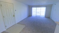 This single-story residential unit features an open living area characterized by neutral-toned walls and a textured ceiling. The main living space is covered in wall-to-wall carpet, while the entryway transitions to tile flooring, creating a practical distinction between the entry and living areas. A large sliding glass door dominates the far wall, allowing abundant natural light to fill the room and providing a view of an outdoor space, likely a patio or balcony. Vertical blinds accompany the sliding door, offering privacy and light control. The layout is straightforward and open, with smooth white walls that offer a blank canvas for personalization. The space appears compact but functional, with clean lines and a simple design typical of apartment-style living. The entry door is positioned to the left, featuring standard hardware, with a light switch panel nearby. Overall, the unit presents a minimalist, move-in-ready environment with practical finishes suited for everyday residential use.