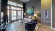 This single-floor residence showcases a modern, open-concept design with impressive soaring ceilings that enhance the sense of space throughout. The living area is anchored by large floor-to-ceiling windows and a glass-paneled door that floods the room with natural light, complemented by wood-look flooring and a neutral color palette accented with bold artwork.

The kitchen is a standout feature, boasting dark cabinetry, stainless steel appliances, a subway tile backsplash, and a granite-topped island with pendant lighting — ideal for both cooking and casual dining, with bar seating provided by teal upholstered chairs.

The bedrooms offer distinct personalities: one features a bold green accent wall paired with a wood-framed bed and walk-in closet access, while another presents a sophisticated gray palette with coordinating navy and teal accents. Each bedroom benefits from ample natural light through large windows with decorative drapery.

The bathrooms are finished with granite vanity tops, dark cabinetry, and tile flooring. One bathroom provides direct access to a walk-in closet, adding practical functionality. Throughout the space, consistent use of teal, navy, and green tones creates a cohesive and polished aesthetic that ties each room together seamlessly.
