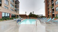 Located at 99 WestEdge Street in Charleston, South Carolina, this outdoor amenity space offers a well-appointed retreat within a modern multi-story residential community. The centerpiece of the area is a generously sized swimming pool with crystal-clear blue water, surrounded by a broad concrete sun deck lined with light blue lounge chairs. An ADA-accessible pool lift ensures the space is welcoming to all residents. Flanking the pool on either side are the community's multi-story buildings, featuring contemporary architecture that blends brick facades with large-paned windows and private balconies. Mature palm trees and lush landscaping add a distinctly Southern coastal character to the setting. Adjacent to the pool area is a spacious open-air courtyard that serves as a social gathering hub. A sleek steel pergola provides partial shade over clusters of weather-resistant sectional sofas and lounge chairs with vibrant turquoise cushions, ideal for outdoor entertaining. String lights are strung between the buildings, lending a warm ambiance to evening gatherings. Together, these outdoor spaces create a cohesive and resort-inspired environment at the heart of this Charleston community.