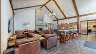 This single-story clubhouse showcases a striking mountain-lodge aesthetic defined by soaring vaulted ceilings with exposed dark wood beams and rich hardwood flooring throughout. The expansive main gathering area serves as the heart of the space, anchored by a dramatic floor-to-ceiling stacked stone fireplace with a reclaimed wood mantel, complemented by multiple seating arrangements featuring leather sofas and armchairs, cowhide rugs, and live-edge wood coffee tables. A large wall-mounted television and curated western-themed artwork enhance the lounge atmosphere.

The full kitchen is outfitted with warm wood cabinetry, a mosaic tile backsplash, stainless steel appliances, and ample counter space. Adjacent to the kitchen, a generously sized stone-clad bar island with a granite countertop accommodates a row of leather-topped bar stools, making it ideal for social gatherings. Multiple pendant chandeliers and recessed lighting illuminate both areas warmly.

A separate game room, accessible through frosted glass sliding doors, provides a more casual recreational space with dark hardwood floors, a pool table, ping-pong table, wall-mounted television, and high-top communal tables, offering a well-rounded amenity space suited for entertainment and community gatherings.