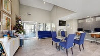Located at 3801 North Belt Line Road in Irving, Texas, this single-floor community space offers a well-designed and functional environment that seamlessly blends a welcoming lounge with practical amenities. The interior is immediately striking thanks to its dramatic vaulted ceilings, which lend an expansive, open feel that belies the footprint of the space.

The primary living and dining area is anchored by bold cobalt blue upholstered seating — including a sofa and a set of coordinating dining chairs arranged around a round bistro-style table — that provides a vibrant contrast against the neutral warm-gray walls and light wood-look tile flooring. Large glass entry doors and sliding windows allow generous natural light to pour in, further brightening the interior.

A wall-mounted flat-screen television and mirrored wall panels add both functionality and visual depth to the lounge area. Adjacent to the main seating zone, a dedicated workstation nook features built-in desk space finished in slate-toned tile accents, equipped with desktop computers and ergonomic seating, making it ideal for business or administrative use.

Framed artwork and a beverage station along one wall complete the space, contributing to an inviting, community-oriented atmosphere. Overall, the space presents a clean, contemporary aesthetic well-suited for gathering, working, and welcoming visitors.