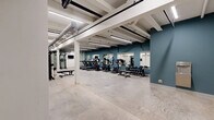 Located at 201 S 12th St in Bessemer City, NC, this fitness center offers a spacious, single-floor layout designed to accommodate a full range of workout needs. The exercise room features an industrial-inspired aesthetic, with exposed structural beams, visible ductwork, and polished concrete flooring that give the space a clean, utilitarian character. Bright linear fluorescent lighting runs throughout the ceiling, ensuring the entire area is well-lit.

The cardio zone is equipped with multiple treadmills, elliptical machines, and stationary bikes — several outfitted with digital displays — arranged in a row alongside large wall-mounted mirrors that enhance the sense of depth and allow users to monitor their form. A dedicated free weights section includes a tiered dumbbell rack with a full range of weights, accompanied by adjustable benches for versatile strength training.

The strength training area features a power rack and additional cable or functional training equipment, with exercise balls and accessories neatly stored nearby. A wall-mounted television is installed for entertainment during workouts. A water fountain is conveniently positioned along one of the teal-accented walls, which provide a bold contrast to the white structural columns throughout the space. A wide doorway connects the fitness center to an adjacent corridor, ensuring easy access and flow.