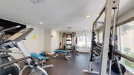Located in El Paso, Texas, this fitness center offers a well-organized, single-room workout space designed to support a comprehensive exercise routine. The room features dark rubber flooring throughout, providing both durability and safety during workouts. Recessed lighting illuminates the space evenly, complemented by several large windows along the walls that allow generous natural light to fill the room and offer views of the surrounding outdoor greenery.

The facility is equipped with a varied selection of strength and cardio equipment. A multi-functional cable trainer serves as a centerpiece for resistance training, accompanied by a full barbell rack loaded with weight plates for compound lifts. A tiered dumbbell rack lines one wall, stocked with a wide range of free weights to accommodate various fitness levels. Adjustable benches in teal upholstery are positioned throughout the floor for versatile use with both free weights and machines.

On the cardio side, elliptical trainers and a rowing machine provide options for low-impact aerobic exercise. A wall-mounted flat-screen television adds an entertainment element to the space. The room also includes a fire extinguisher and clearly marked exit signage, ensuring safety compliance. The overall ambiance is clean, functional, and practical, making it a well-rounded facility for daily fitness needs.
