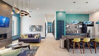 Located in Scott, Louisiana, this single-floor clubhouse offers a thoughtfully designed open-concept layout that seamlessly combines a fully equipped kitchen with a spacious living area. The interior design makes a bold statement through vibrant teal accent walls, high ceilings, and light wood-tone flooring that runs throughout the space.

The living area serves as the social centerpiece, featuring multiple plush navy sofas arranged around a sculptural wood coffee table atop a richly patterned area rug. A dramatic floor-to-ceiling stone tile fireplace wall anchors the room, complete with a wall-mounted flat-screen TV and a striking multi-arm sculptural chandelier overhead. Large windows with white plantation shutters flood the space with natural light and offer views of the surrounding outdoor area.

The kitchen is appointed with deep navy cabinetry, a textured gray tile backsplash, dark stone countertops with bold veining, open shelving, and a stainless steel side-by-side refrigerator. A kitchen island with a richly marbled countertop provides additional workspace and casual seating. A compact dining nook with a white table and modern chairs is nestled near the bright windowed corner. Curated abstract artwork displayed throughout adds a layer of sophistication to this well-appointed communal space.