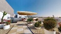 Located along North Coast Highway in Oceanside, California, this rooftop terrace offers a well-appointed outdoor retreat with panoramic views of the surrounding coastal landscape. The space is defined by a striking checkered tile floor, alternating between matte and textured stone finishes, which adds a bold visual rhythm throughout the terrace. Thoughtfully curated desert-inspired landscaping — including agave, ornamental grasses, and sculptural shrubs arranged in large white planters with gravel beds — gives the space a refined, resort-like atmosphere. Multiple seating zones are distributed across the terrace to accommodate a variety of uses. A generous lounge area features plush teal sectional sofas and round terrazzo side tables, shaded by signature fringed yellow cantilever umbrellas. A dedicated dining area includes sleek tables paired with vibrant red chairs, also sheltered under matching oversized umbrellas. Additional amenities include an outdoor kitchen with a built-in grill, a shuffleboard table for recreation, and a wall-mounted television for outdoor entertainment. The open parapet walls allow unobstructed sightlines across the Oceanside skyline and toward the Pacific horizon, making this terrace an exceptional space for gatherings, relaxation, and al fresco dining under the Southern California sun.